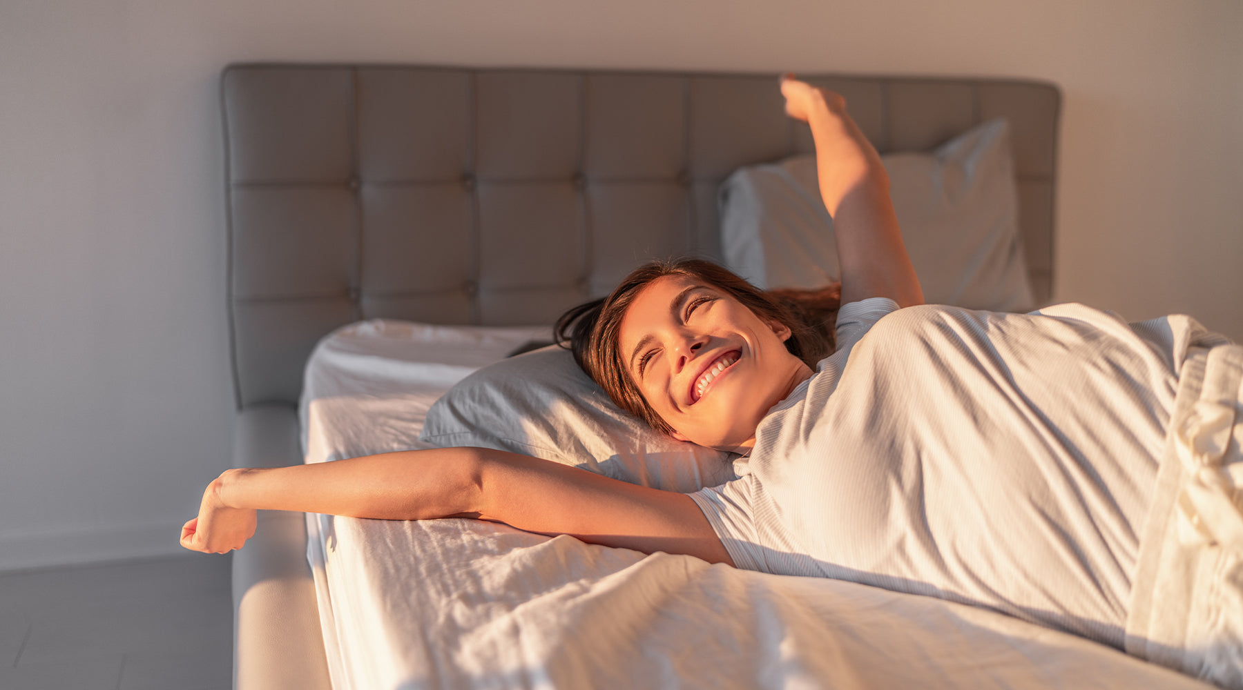 Smilende kvinde strækker sig i sengen med morgensolen i ansigtet